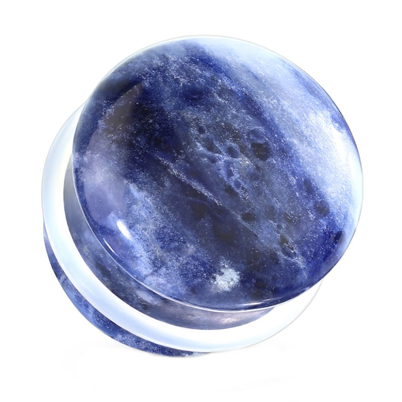A round, polished blue sodalite stone plug earring with white streaks and dark patches holding a clear o-ring around it, displayed from a slightly elevated angle. The surface is smooth and glossy, highlighting its natural patterns and it's pictured on a white background.